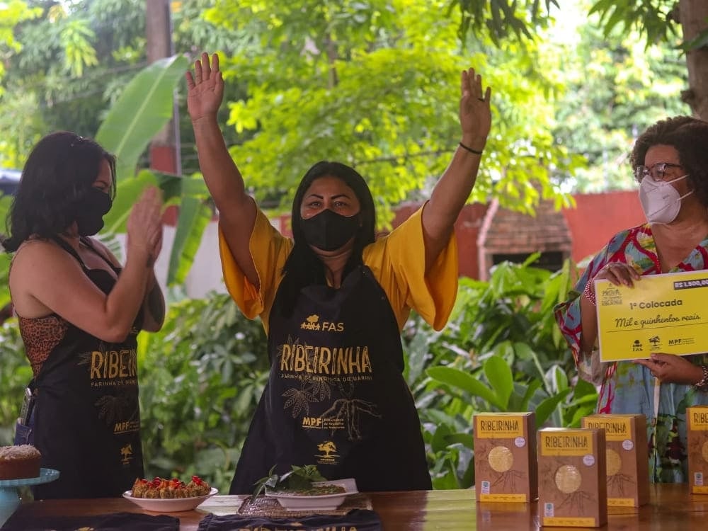 Une femme portant un tablier noir avec le logo FAS Ribeirinha lève les mains de joie, aux côtés d'autres personnes célébrant le programme de la Fondation Amazonie Durable. Une femme portant un tablier noir avec le logo FAS Ribeirinha lève les mains de joie, aux côtés d'autres personnes célébrant le programme de la Fondation Amazonie Durable.
