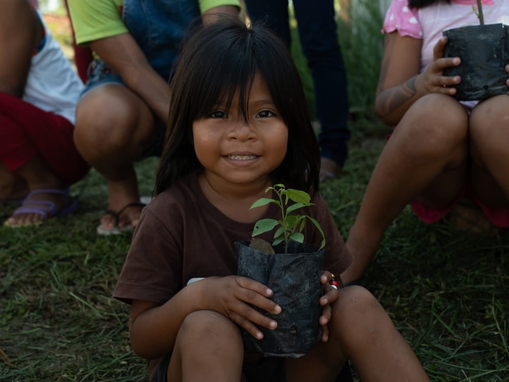 Uma criança sorridente de uma comunidade ribeirinha da Amazônia sentada na grama segurando uma pequena planta no vaso. Uma criança sorridente de uma comunidade ribeirinha da Amazônia sentada na grama segurando uma pequena planta no vaso.