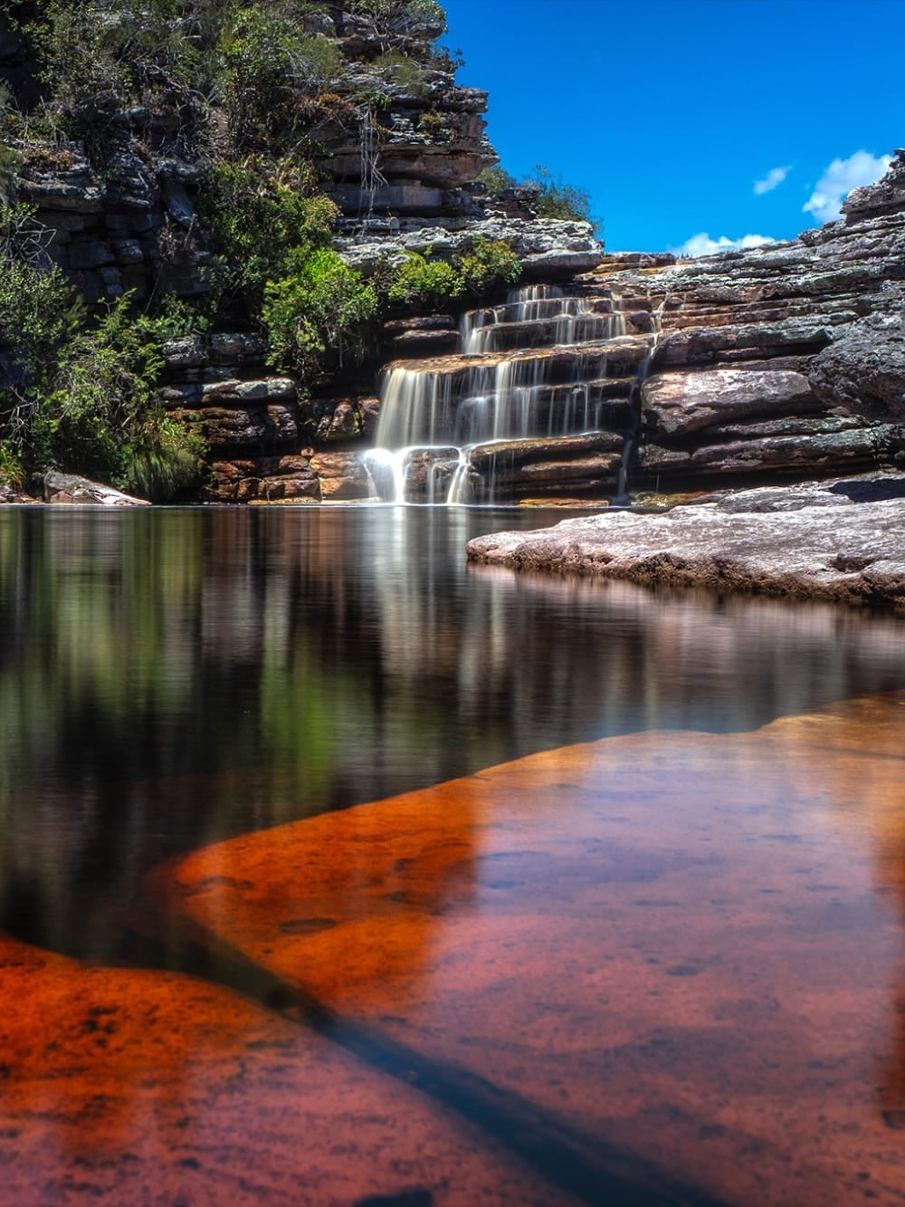 Uma cachoeira em cascata desaguando em um poço natural calmo com rochas avermelhadas no Parque Nacional da Chapada Diamantina. Uma cachoeira em cascata desaguando em um poço natural calmo com rochas avermelhadas no Parque Nacional da Chapada Diamantina.