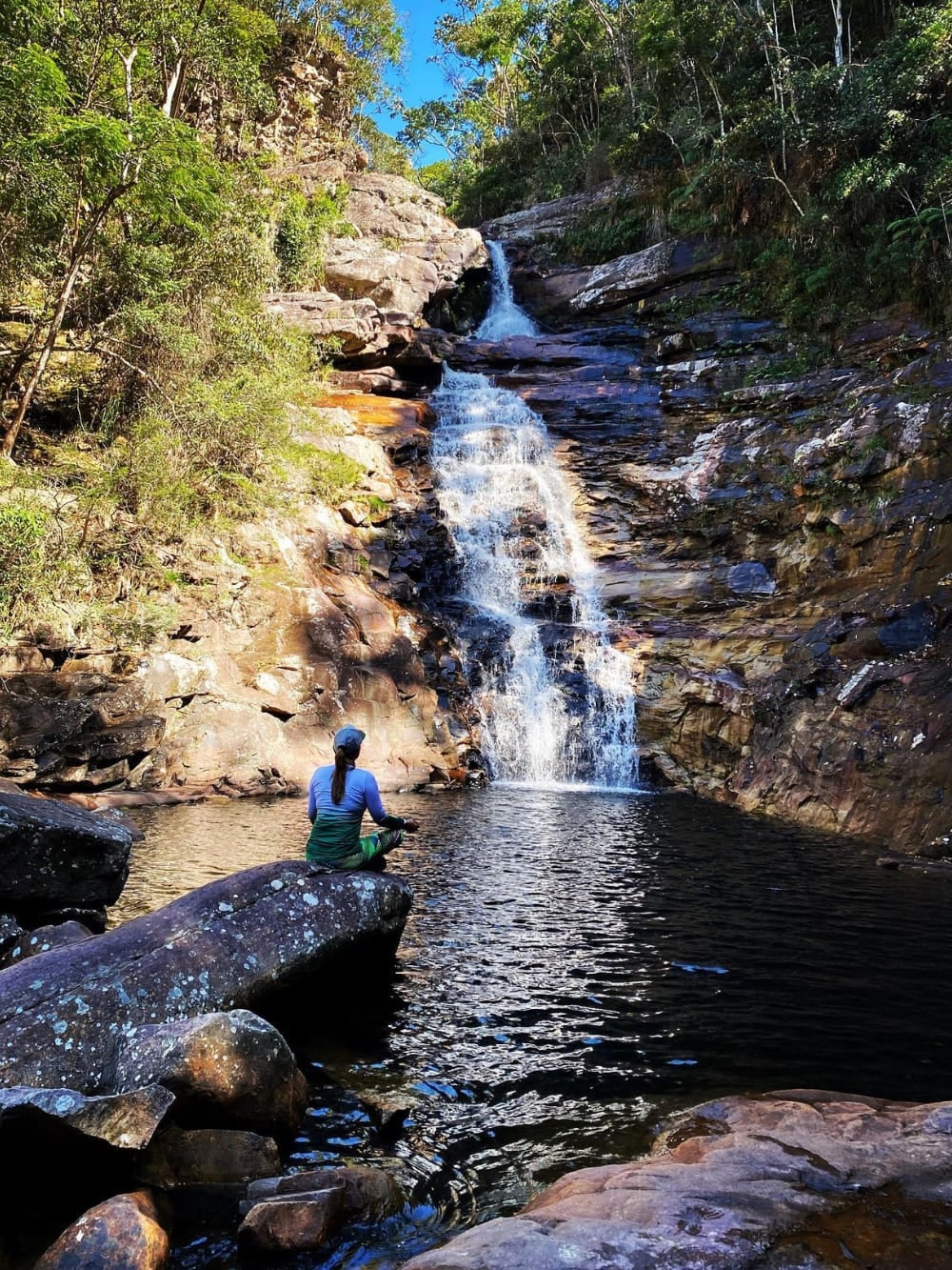 Uma mulher sentada em uma rocha perto do poço natural calmo da Cachoeira do Funil, uma queda de 20 metros cercada por vegetação verde densa. Uma mulher sentada em uma rocha perto do poço natural calmo da Cachoeira do Funil, uma queda de 20 metros cercada por vegetação verde densa.