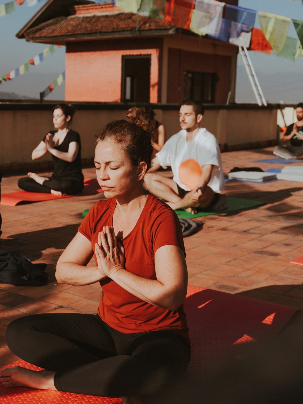 Mila Ricetti en Lorenzo die yoga beoefenen en mediteren met een groep op een zonovergoten dakterras versierd met gebedsvlaggen tijdens een retraite in Nepal. Mila Ricetti en Lorenzo die yoga beoefenen en mediteren met een groep op een zonovergoten dakterras versierd met gebedsvlaggen tijdens een retraite in Nepal.