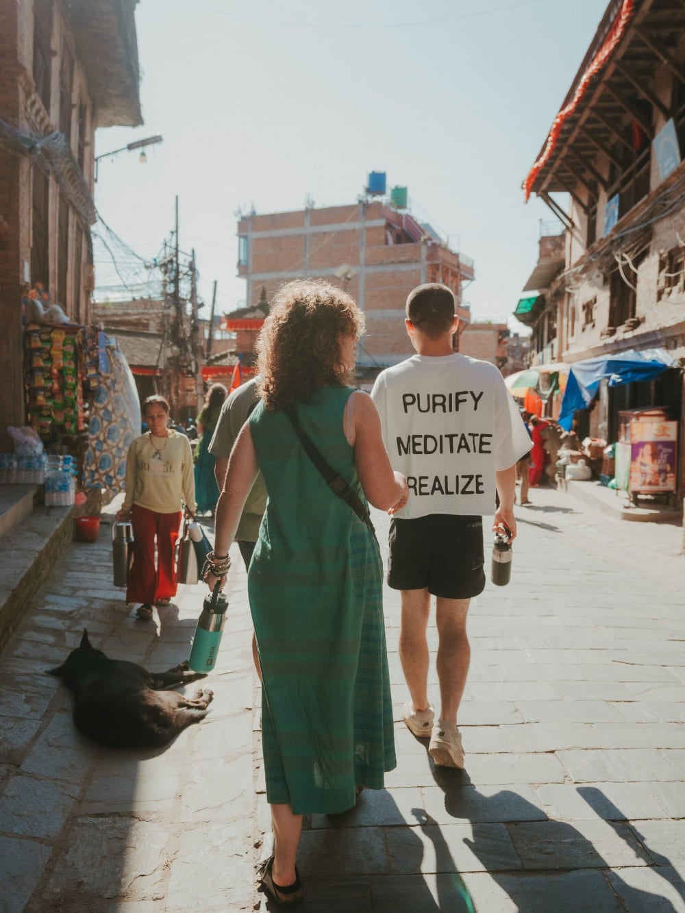 Mila en Lorenzo lopen zij aan zij door een zonnige straat in Nepal, waarbij Lorenzo een T-shirt draagt met de tekst 'purify, meditate, realize'. Mila en Lorenzo lopen zij aan zij door een zonnige straat in Nepal, waarbij Lorenzo een T-shirt draagt met de tekst 'purify, meditate, realize'.