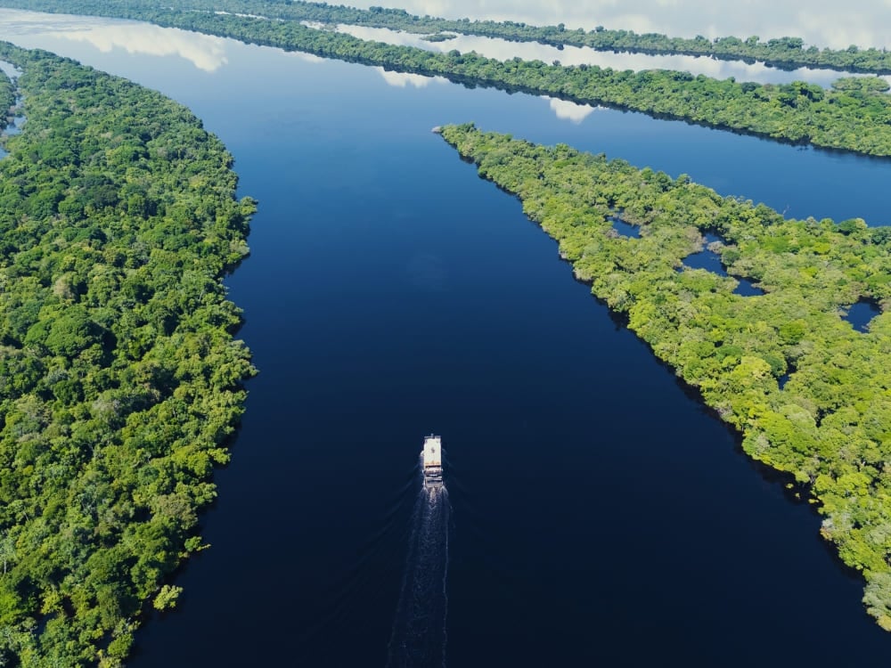 Um barco de cruzeiro fluvial de vários andares viaja ao longo de um rio escuro e calmo, cercado por densas ilhas verdes de floresta tropical que refletem o céu. Um barco de cruzeiro fluvial de vários andares viaja ao longo de um rio escuro e calmo, cercado por densas ilhas verdes de floresta tropical que refletem o céu.