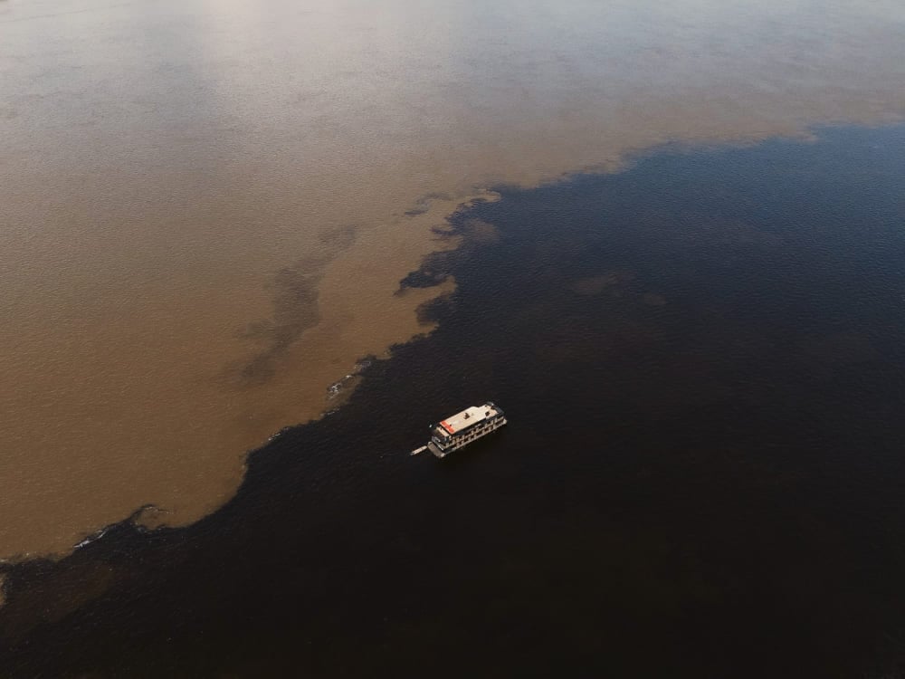 Vista aérea de um barco navegando pelo Encontro das Águas na Amazônia, mostrando os distintos rios escuro e barrento fluindo lado a lado. Vista aérea de um barco navegando pelo Encontro das Águas na Amazônia, mostrando os distintos rios escuro e barrento fluindo lado a lado.