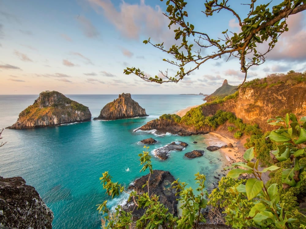 As icônicas formações rochosas do Morro dos Dois Irmãos se erguem do oceano turquesa ao longo de uma exuberante praia cercada por penhascos em Fernando de Noronha. As icônicas formações rochosas do Morro dos Dois Irmãos se erguem do oceano turquesa ao longo de uma exuberante praia cercada por penhascos em Fernando de Noronha.