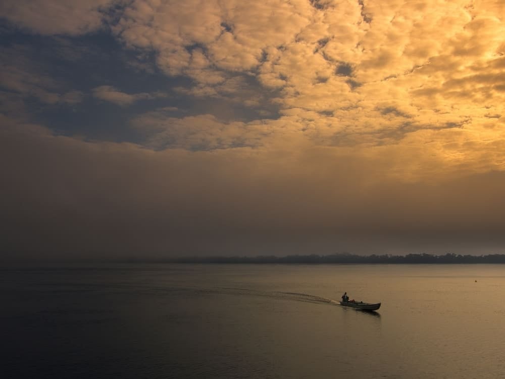 Een kleine gemotoriseerde kano vaart over de brede rivier tijdens de zonsopgang onder een bewolkte lucht; ideaal voor wie de Amazone bezoeken wil. Een kleine gemotoriseerde kano vaart over de brede rivier tijdens de zonsopgang onder een bewolkte lucht; ideaal voor wie de Amazone bezoeken wil.