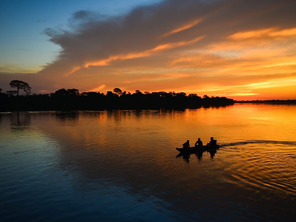 Silhouet van drie mensen in een kleine kano die varen over de rivier tegen een feloranje zonsondergang. Silhouet van drie mensen in een kleine kano die varen over de rivier tegen een feloranje zonsondergang.