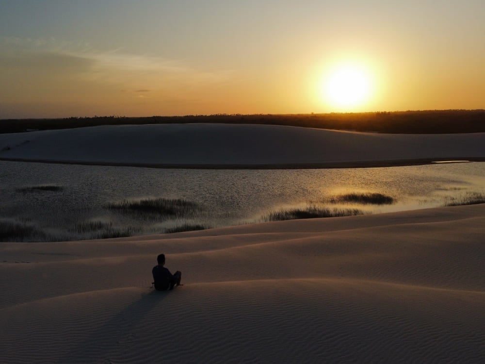 Silhouette einer Person, die auf einer Sanddüne sitzt und einen goldenen Sonnenuntergang über den Lagunen der Lençóis Maranhenses beobachtet. Silhouette einer Person, die auf einer Sanddüne sitzt und einen goldenen Sonnenuntergang über den Lagunen der Lençóis Maranhenses beobachtet.