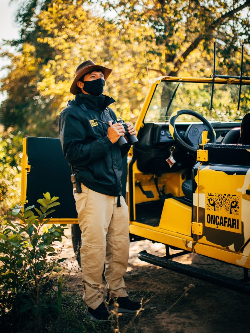 Um guia do projeto Onçafari usando máscara segura binóculos enquanto está ao lado de um veículo amarelo de safári no Pantanal. Um guia do projeto Onçafari usando máscara segura binóculos enquanto está ao lado de um veículo amarelo de safári no Pantanal.