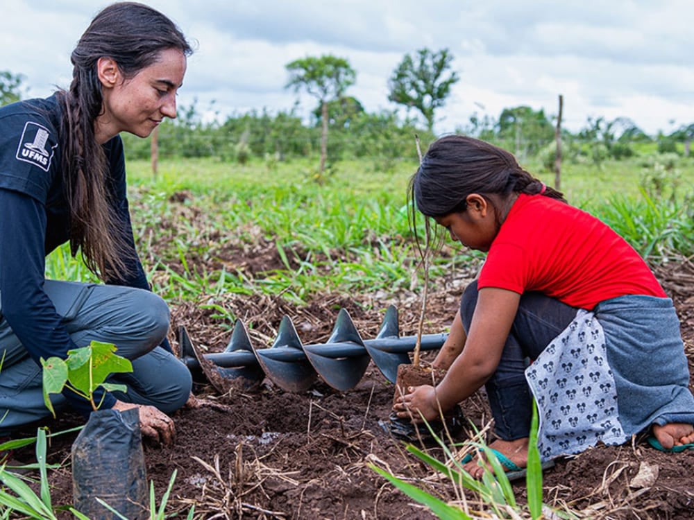 Uma mulher do projeto SOS Pantanal e uma jovem garota local usam um perfurador de solo para plantar uma muda de árvore juntas. Uma mulher do projeto SOS Pantanal e uma jovem garota local usam um perfurador de solo para plantar uma muda de árvore juntas.