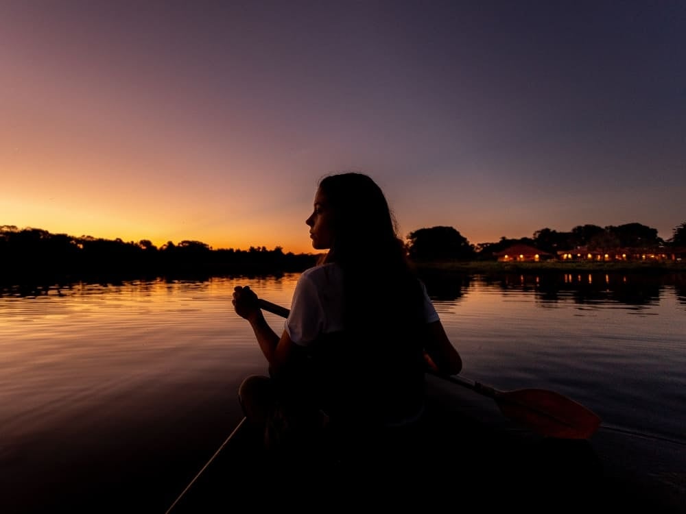 Silhouette einer Person, die bei einem friedlichen Sonnenuntergang in einem Kanu auf dem ruhigen Wasser eines Flusses paddelt. Silhouette einer Person, die bei einem friedlichen Sonnenuntergang in einem Kanu auf dem ruhigen Wasser eines Flusses paddelt.