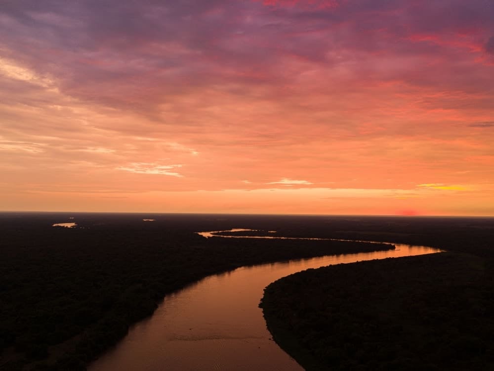 Luftaufnahme eines sich windenden Flusses, der durch die weiten Feuchtgebiete fließt und den rosa-orangefarbenen Abendhimmel widerspiegelt. Luftaufnahme eines sich windenden Flusses, der durch die weiten Feuchtgebiete fließt und den rosa-orangefarbenen Abendhimmel widerspiegelt.