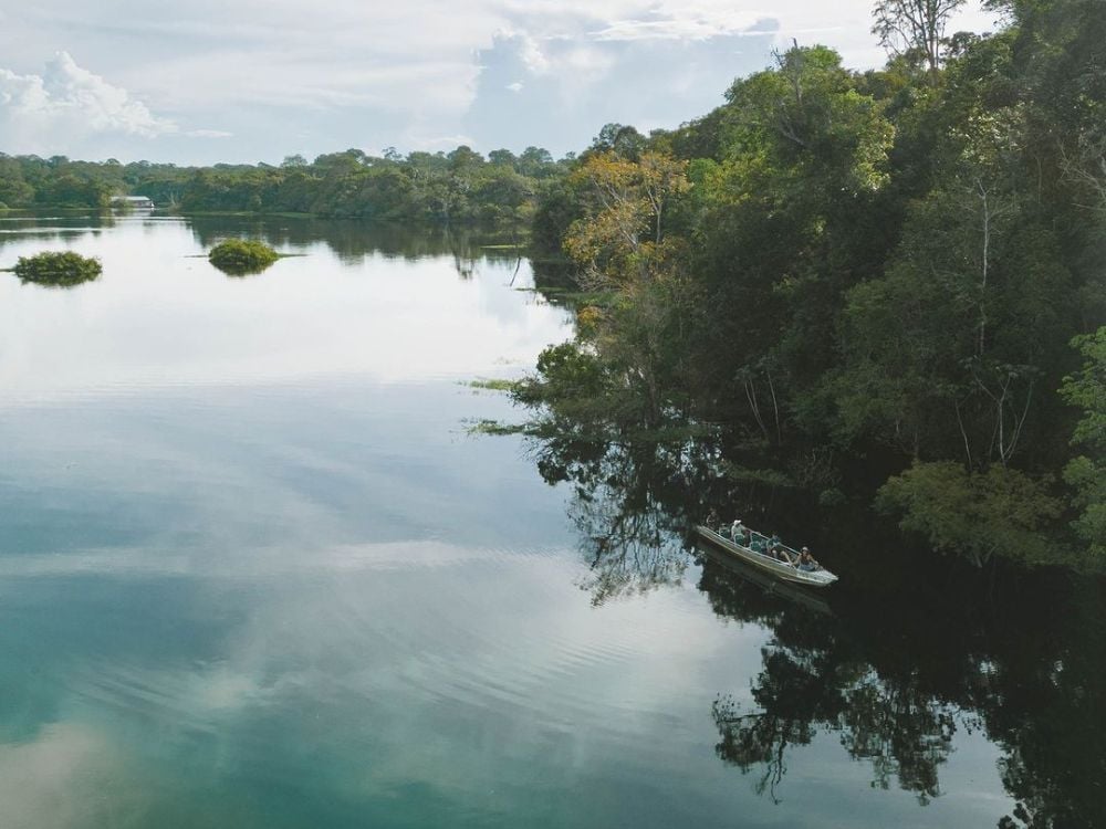 Pequeno barco navegando à distância nas águas calmas e reflexivas do Rio Mamori, cercado pela vegetação verde da selva. Pequeno barco navegando à distância nas águas calmas e reflexivas do Rio Mamori, cercado pela vegetação verde da selva.