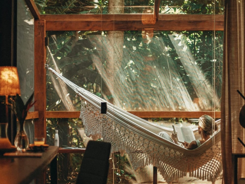Une personne se relaxant dans un hamac à l'intérieur d'une chambre sophistiquée avec de grandes baies vitrées donnant sur la jungle. Une personne se relaxant dans un hamac à l'intérieur d'une chambre sophistiquée avec de grandes baies vitrées donnant sur la jungle.