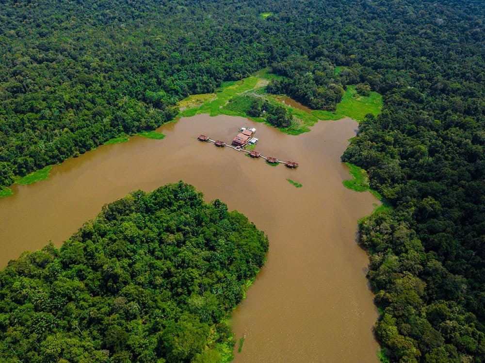 Vue aérienne d'un éco-lodge avec des structures flottantes sur une rivière sinueuse entièrement entourée d'une forêt dense. Vue aérienne d'un éco-lodge avec des structures flottantes sur une rivière sinueuse entièrement entourée d'une forêt dense.