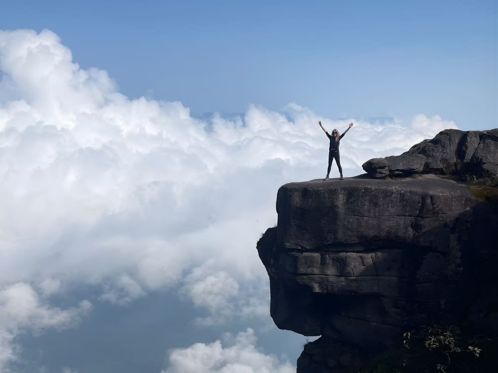 Une femme voyageant seule, debout au bord d’une haute falaise au-dessus des nuages, lève les bras en signe de victoire lors d’une aventure en montagne au Brésil. Une femme voyageant seule, debout au bord d’une haute falaise au-dessus des nuages, lève les bras en signe de victoire lors d’une aventure en montagne au Brésil.