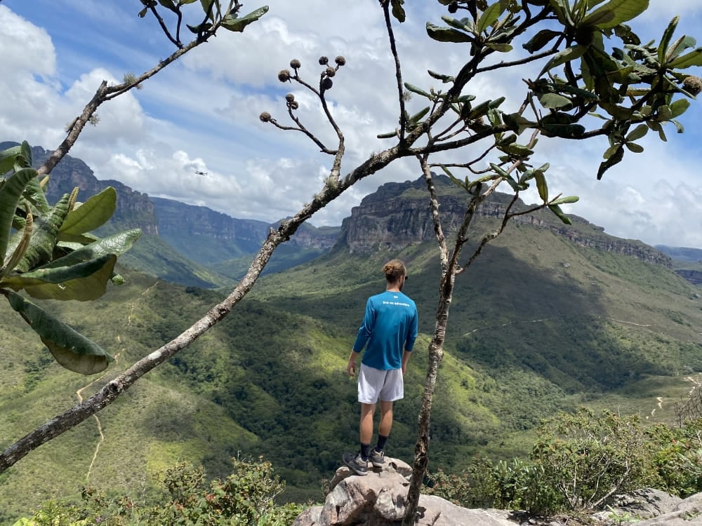 Um trilheiro em pé na beira de uma rocha, emoldurado por galhos de árvores, observando o vale verde em direção ao topo plano do Morro do Castelo. Um trilheiro em pé na beira de uma rocha, emoldurado por galhos de árvores, observando o vale verde em direção ao topo plano do Morro do Castelo.