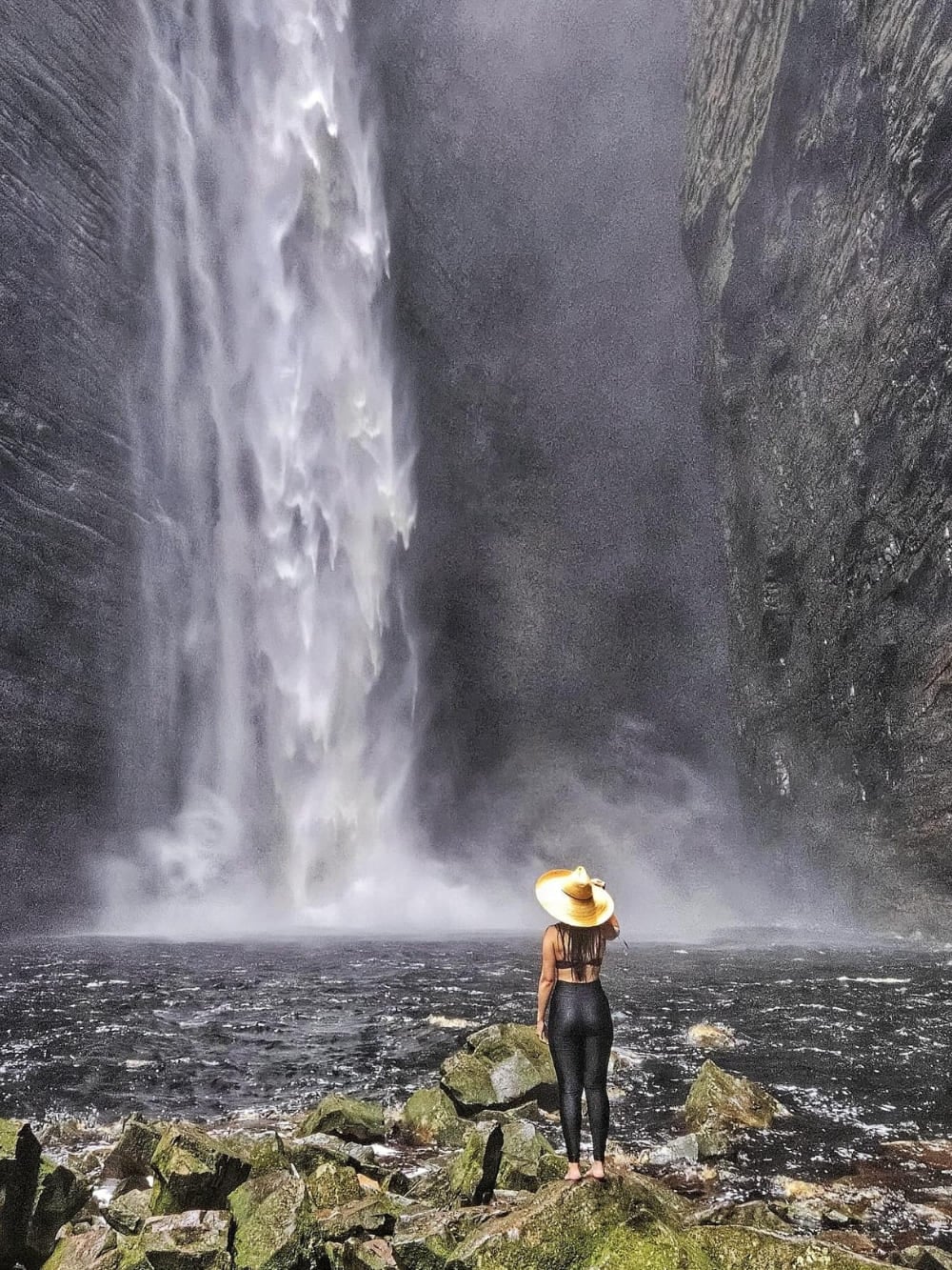 Een wandelaar met hoed staat aan de rand van een donker zwembad en kijkt omhoog naar de dramatische verticale val van 100 meter van de Fumacinha-waterval in een smalle kloof. Een wandelaar met hoed staat aan de rand van een donker zwembad en kijkt omhoog naar de dramatische verticale val van 100 meter van de Fumacinha-waterval in een smalle kloof.