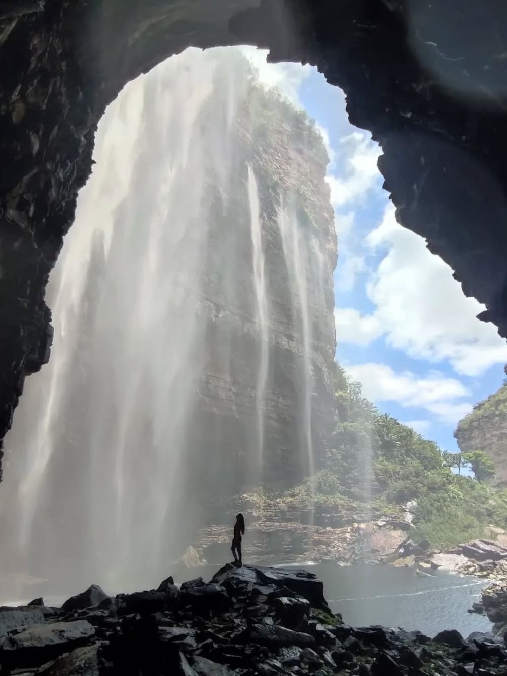 Een silhouet van een persoon die op rotsen staat bij de ingang van een donkere grot en uitkijkt op de ruige kliffen van 100 meter van de Herculano-waterval. Een silhouet van een persoon die op rotsen staat bij de ingang van een donkere grot en uitkijkt op de ruige kliffen van 100 meter van de Herculano-waterval.