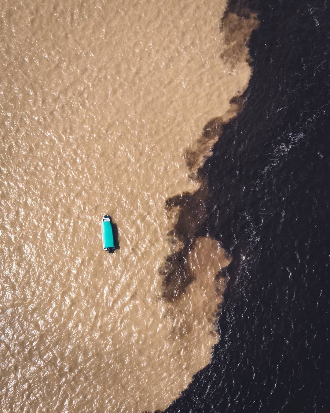 Um barco pequeno, dois rios que teimam em não se misturar. Vista aérea de um pequeno barco com cobertura verde-água navegando no Encontro das Águas, onde as águas barrentas do Solimões encontram as águas escuras do Rio Negro em um cruzeiro pela Amazônia no Brasil