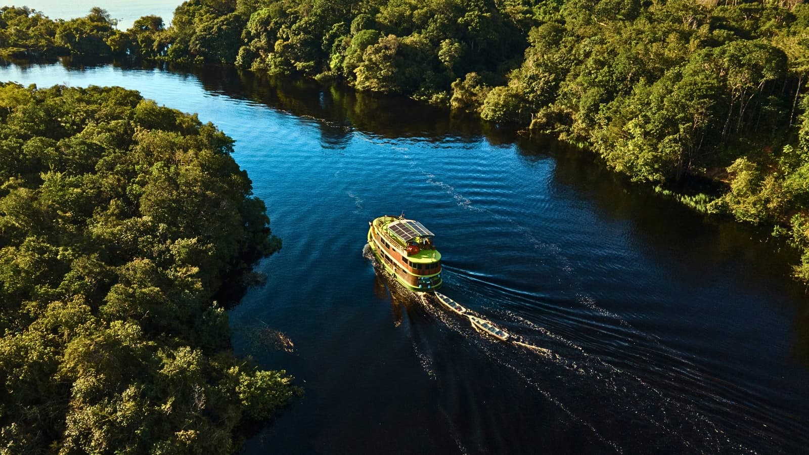 O barco abre caminho entre a floresta como se sempre fosse dali. Vista aérea de um barco de cruzeiro fluvial na Amazônia com painéis solares, navegando um rio de águas negras com uma pequena embarcação rebocada, ladeado por densa floresta tropical
