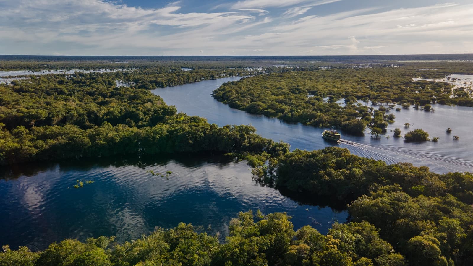 Hier gibt der Fluss die Richtung vor, und der Wald schaut einfach zu. Luftaufnahme eines Kreuzfahrtschiffs auf einem gewundenen Schwarzwasserfluss inmitten überschwemmten Regenwaldes beim Amazon Jungle Cruise Brazil, mit baumbedecktem Horizont unter leicht bewölktem Himmel