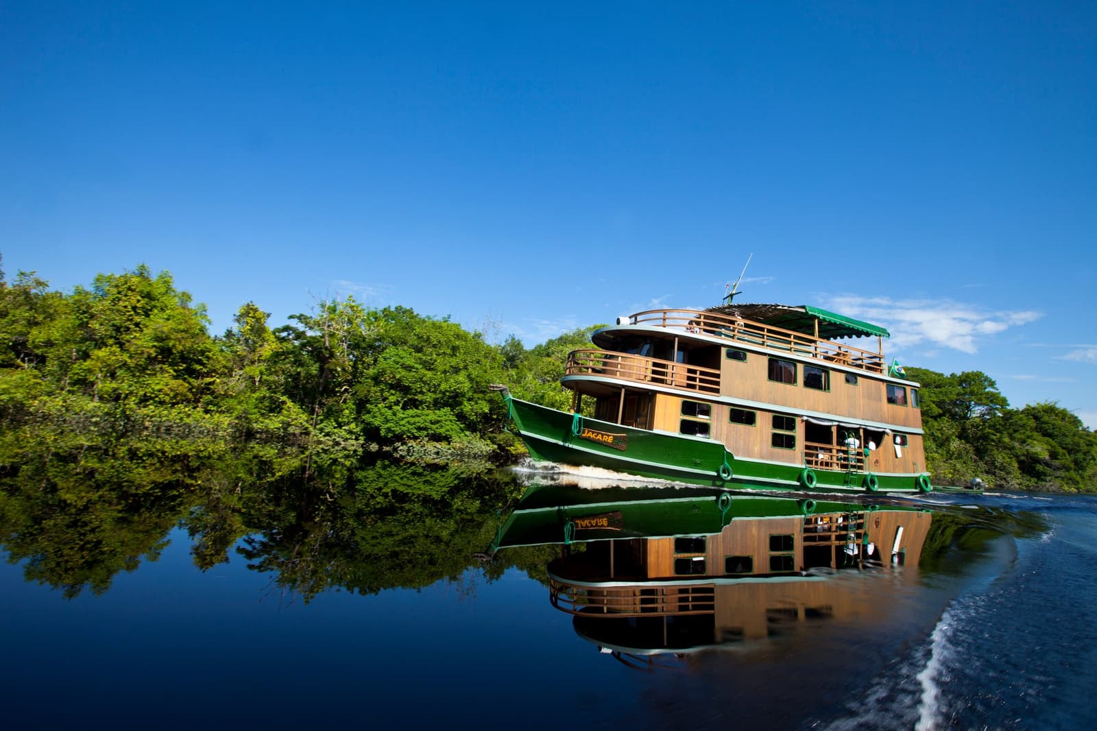 Das schwimmende Zuhause für ein paar Tage tiefer Amazonas-Stille. Das hölzerne Hausboot Jacaré gleitet auf einem Schwarzwasserkanal durch den brasilianischen Amazonas-Dschungel-Kreuzfahrt, der grüne Rumpf spiegelt sich perfekt im stillen Wasser, begleitet von dichtem Regenwald unter blauem Himmel