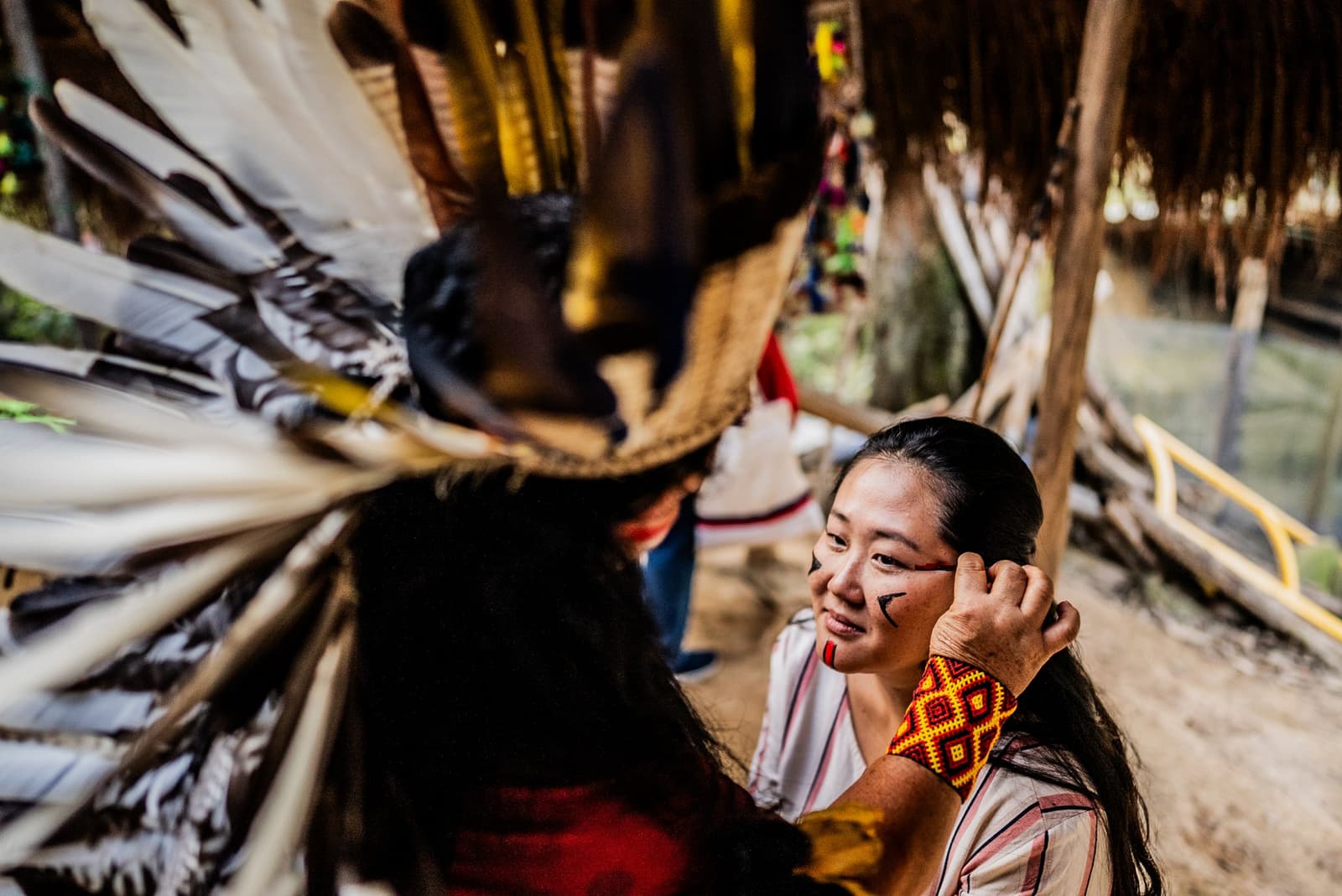 The lines drawn here carry stories older than any map. A smiling visitor receives traditional geometric face paint from an Indigenous community member wearing a large feathered headdress during an Amazon River Cruise Brazil cultural encounter
