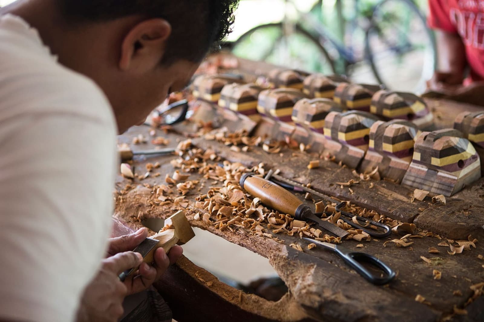 Des années de pratique, quelques ciseaux, et des pièces qu'on embarquerait toutes dans ses bagages. Un artisan sculpte à la main une petite pièce de bois sur un établi couvert de copeaux, tandis que des figurines en bois lamellé multicolore sont alignées à côté de ses outils