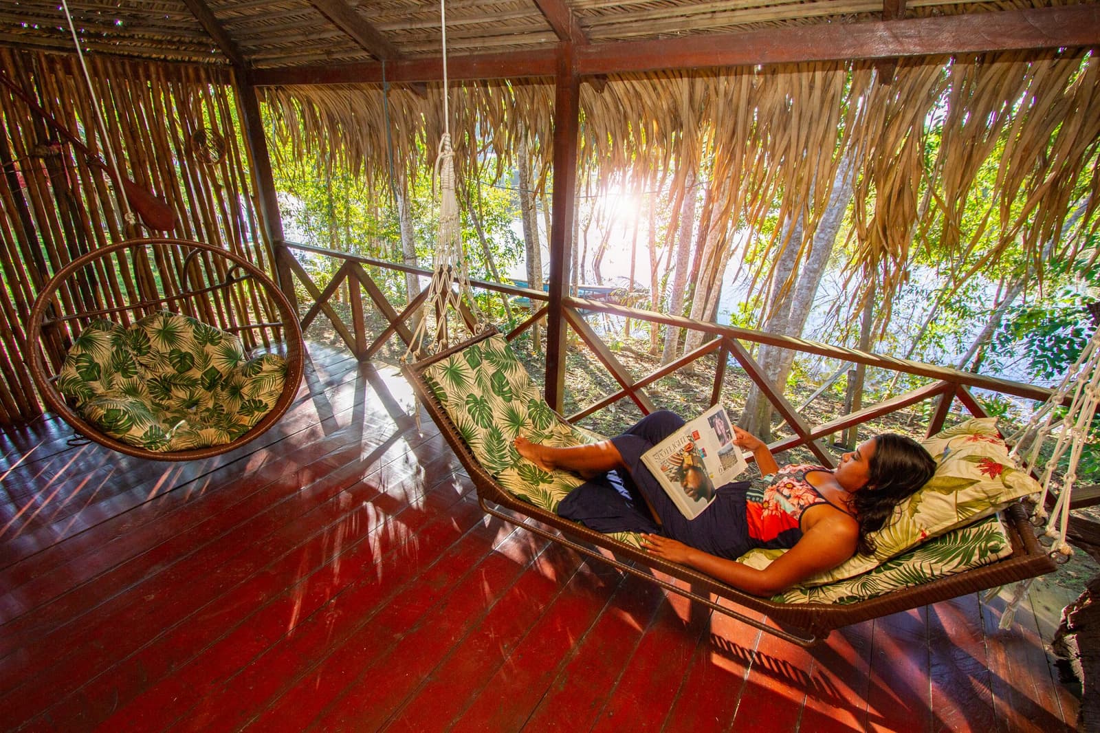 Some afternoons in the Amazon, a magazine and a hammock are all the agenda you need. A guest reclines on a cushioned hammock swing reading a magazine on the thatched open-air terrace of the Amazon Turtle Lodge, with sunlit rainforest trees visible beyond the wooden railing