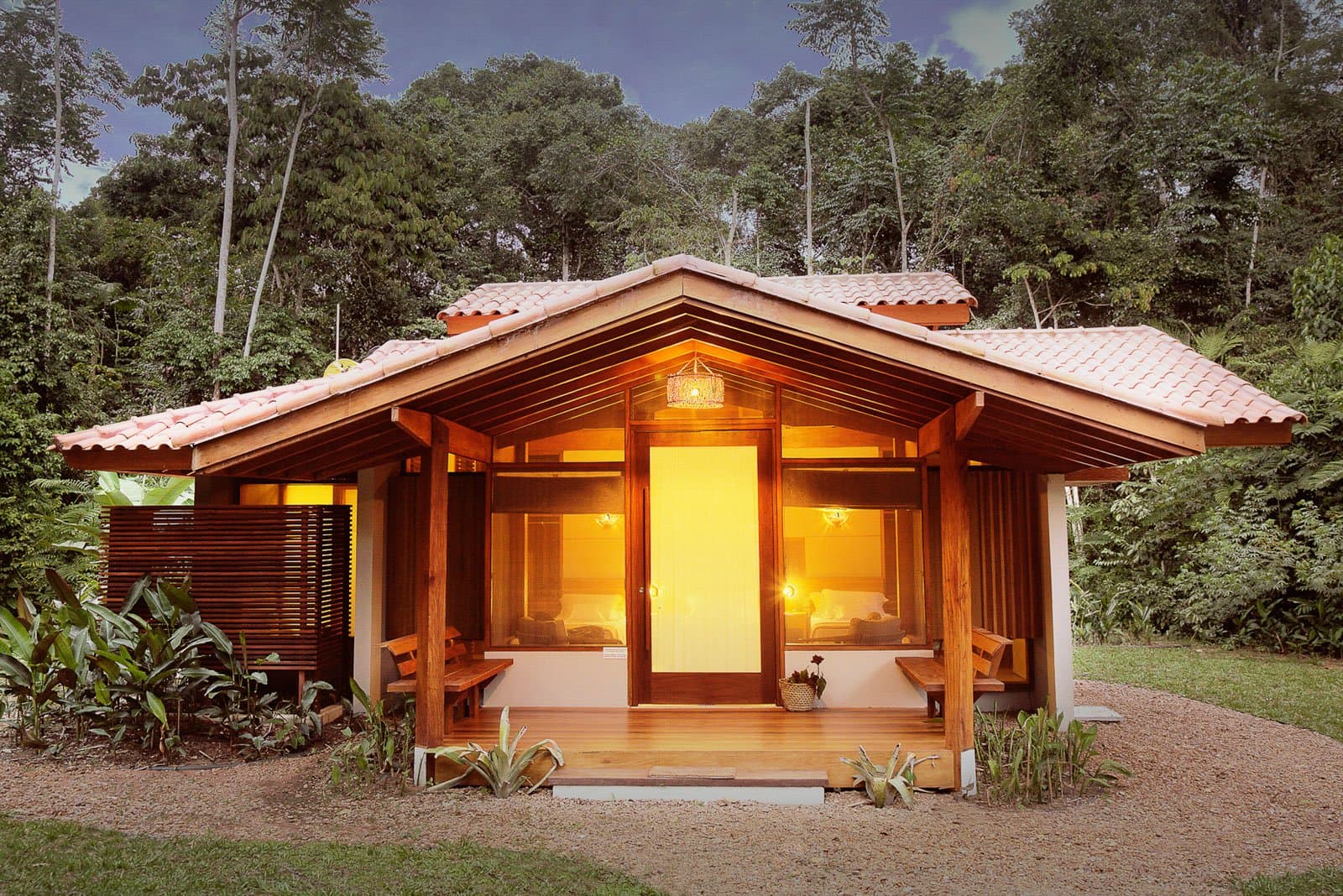 Cosy enough to make you forget the 4 AM alarm. Almost. Photo: Samuel Melim (c) A warm-lit wooden bungalow with terracotta roof tiles, a covered front veranda with timber benches, and large glass doors glowing amber at dusk, set against dense Amazon rainforest — part of the birdwatching in the Amazon rainforest experience at Cristalino Jungle Lodge