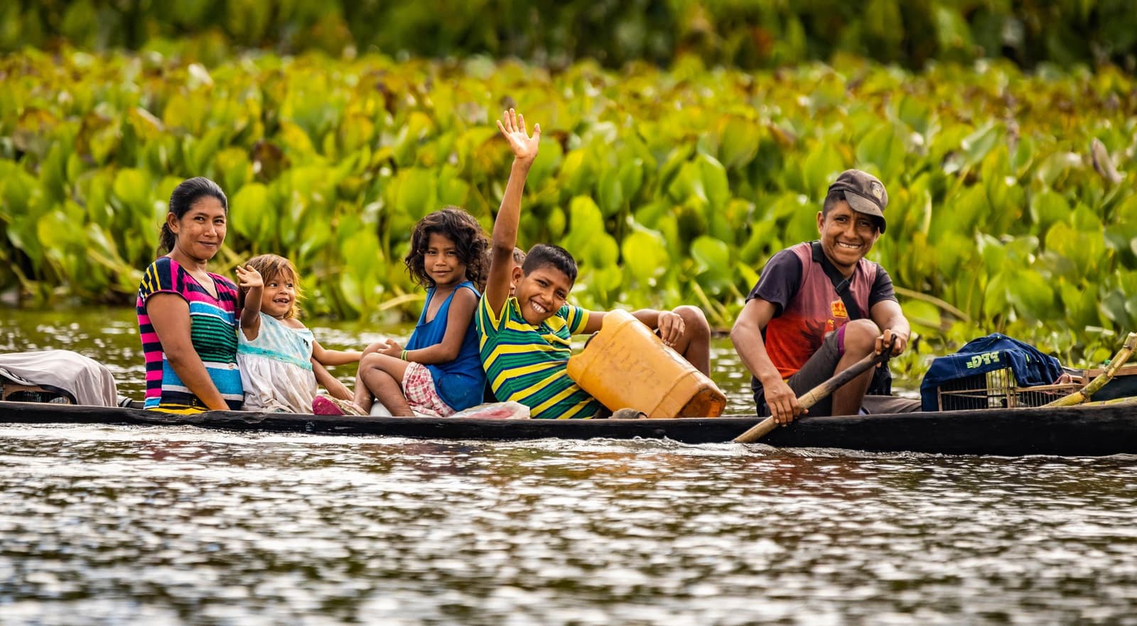 Toda viagem pela Amazônia tem um momento assim, guarde o seu. Uma família sorridente de cinco pessoas rema em uma canoa de madeira por um rio durante um passeio pela selva amazônica brasileira, com um menino acenando animado e vegetação aquática exuberante ao fundo