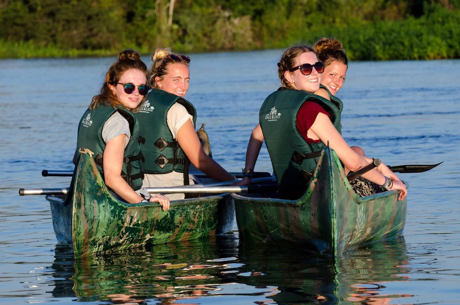 Gute Gesellschaft auf ruhigem Wasser, mehr braucht es nicht. Vier lachende Frauen in grünen Schwimmwesten paddeln während einer günstigen Pantanal-Tour in einem Kanu auf einem ruhigen Fluss, dichter grüner Wald am gegenüberliegenden Ufer