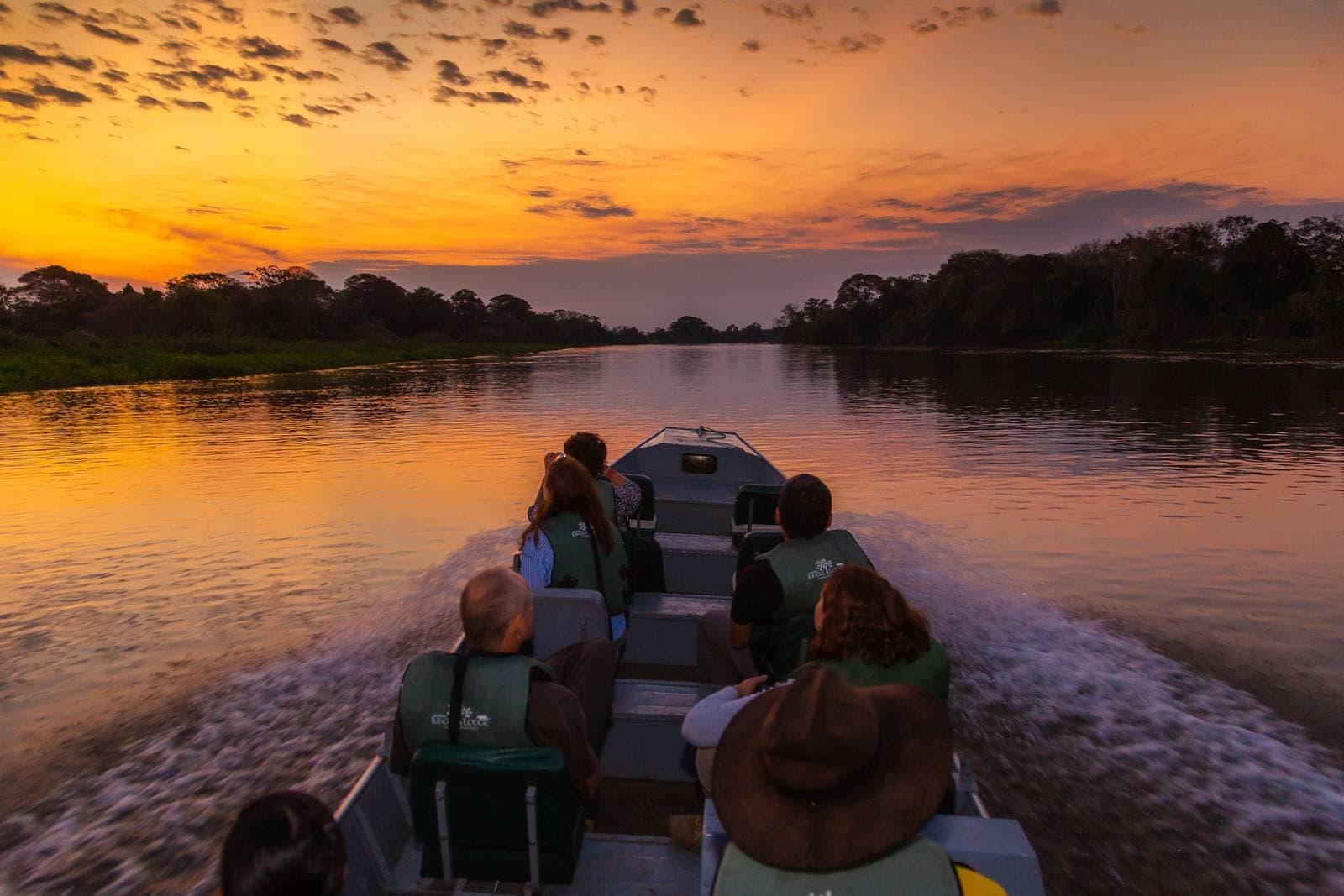 Sonnenuntergang mit Fahrtwind, so sollte jeder Abend enden. Reisende in Schwimmwesten auf einem Motorboot, das bei einem intensiv orangefarbenen Sonnenuntergang einen ruhigen Flusskanal im Pantanal entlangfährt, umgeben von Waldsилуetten