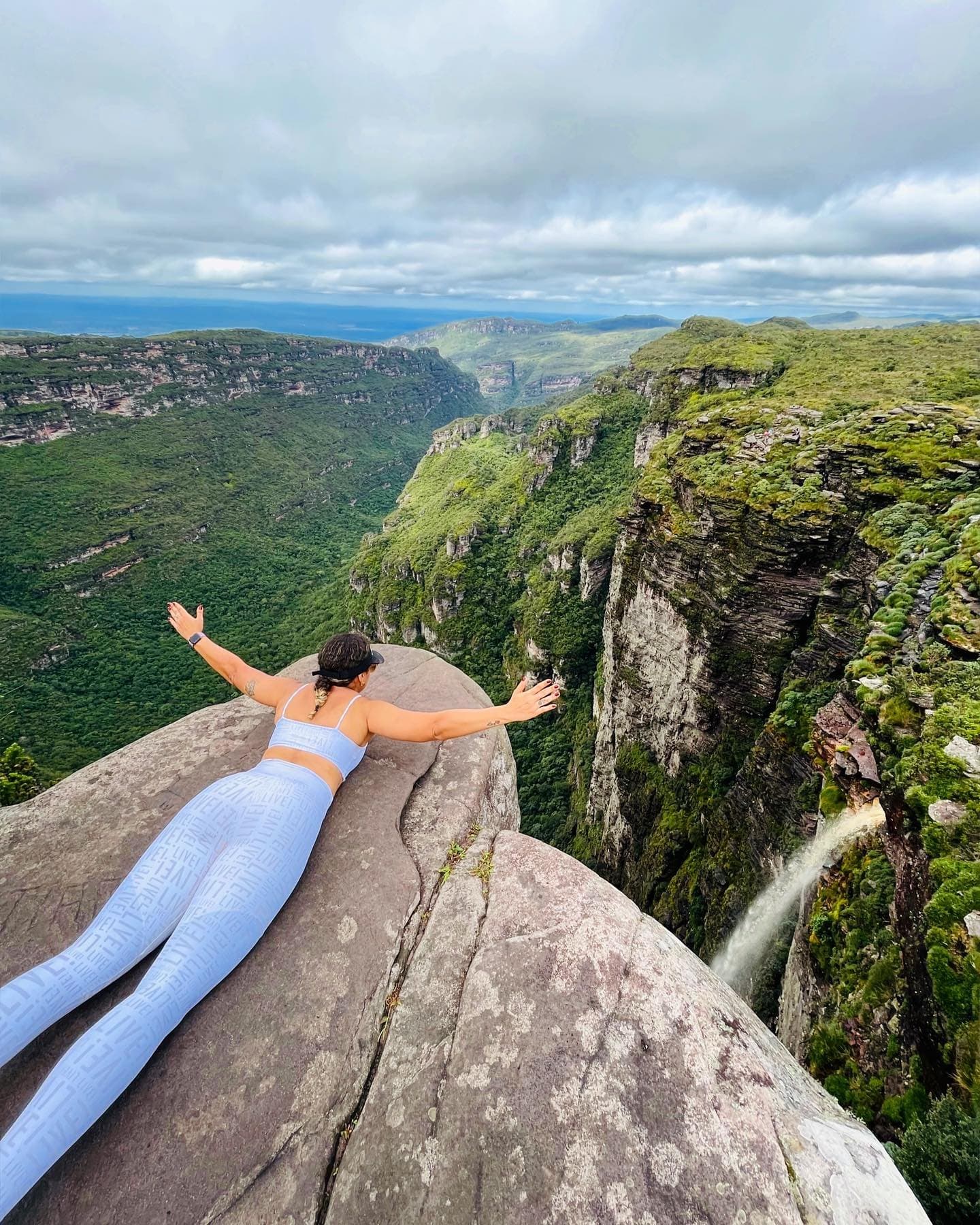 Les bras grands ouverts sur le vide, la cascade en fond sonore. Une femme est allongée bras écartés sur un rocher au bord d'un canyon profond lors du Chapada Diamantina Tour, une cascade dévalant la falaise sur la droite et des plateaux boisés s'étendant jusqu'à l'horizon