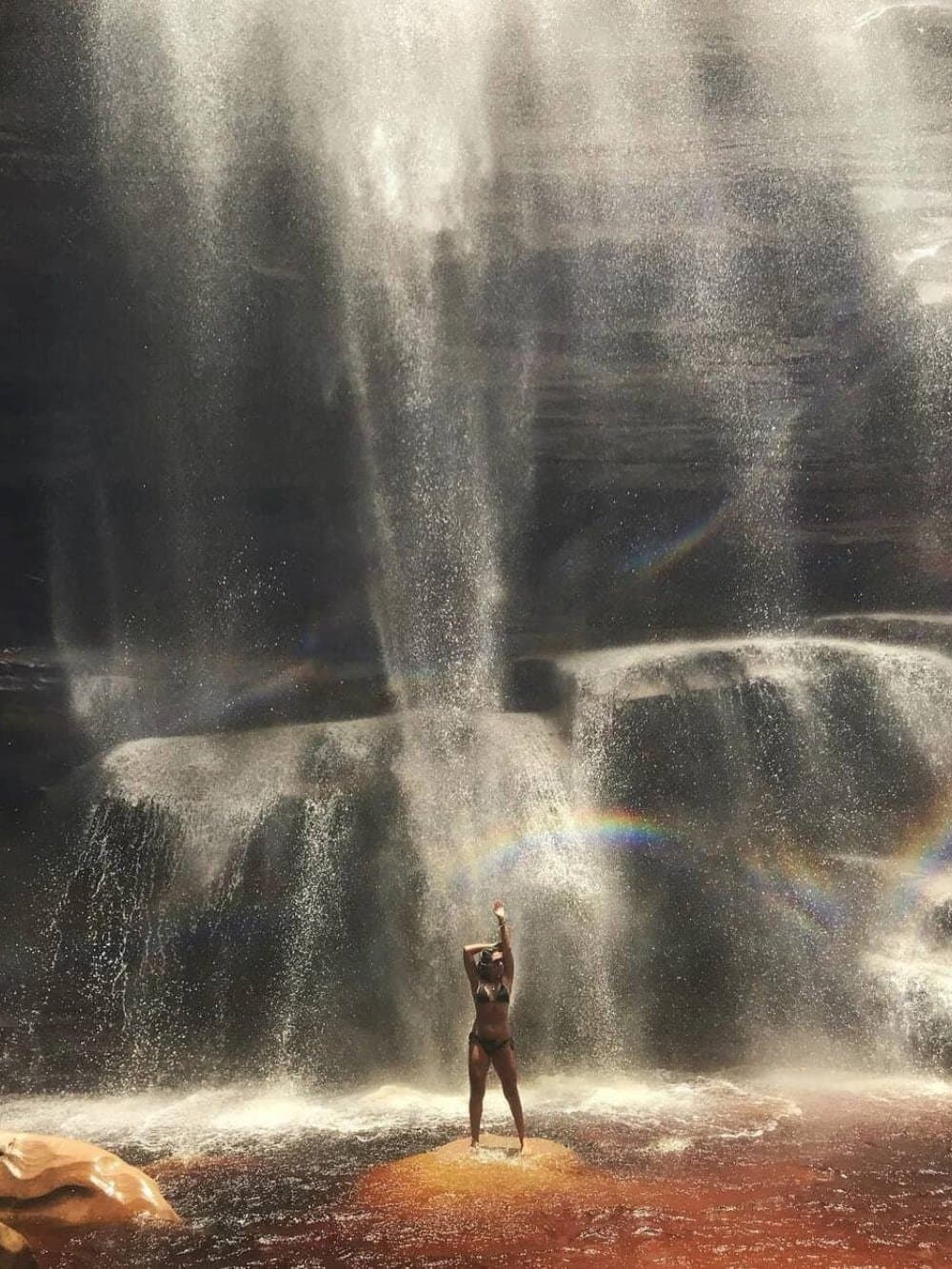 L'arc-en-ciel était là, il ne restait qu'à lever les bras. Une visiteuse en bikini, les bras levés, se tient sur un rocher sous une grande cascade lors du circuit Chapada Diamantina, avec un arc-en-ciel vif traversant les embruns au premier plan