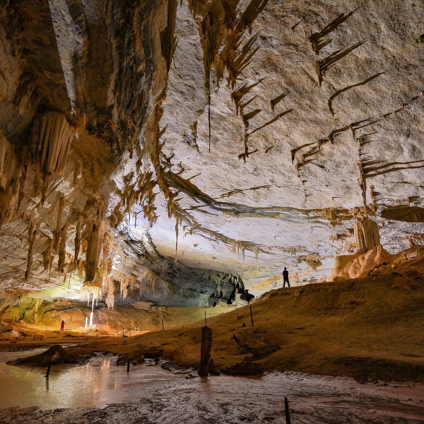 Millionen Jahre Geduld, in Stein geschrieben. Zwei winzige menschliche Silhouetten stehen in der riesigen, kathedralenartigen Halle einer Kalksteinhöhle auf einer Chapada-Diamantina-Reise, überragt von Stalaktiten an der gewölbten Decke und einem flachen Spiegelpool auf dem Höhlenboden