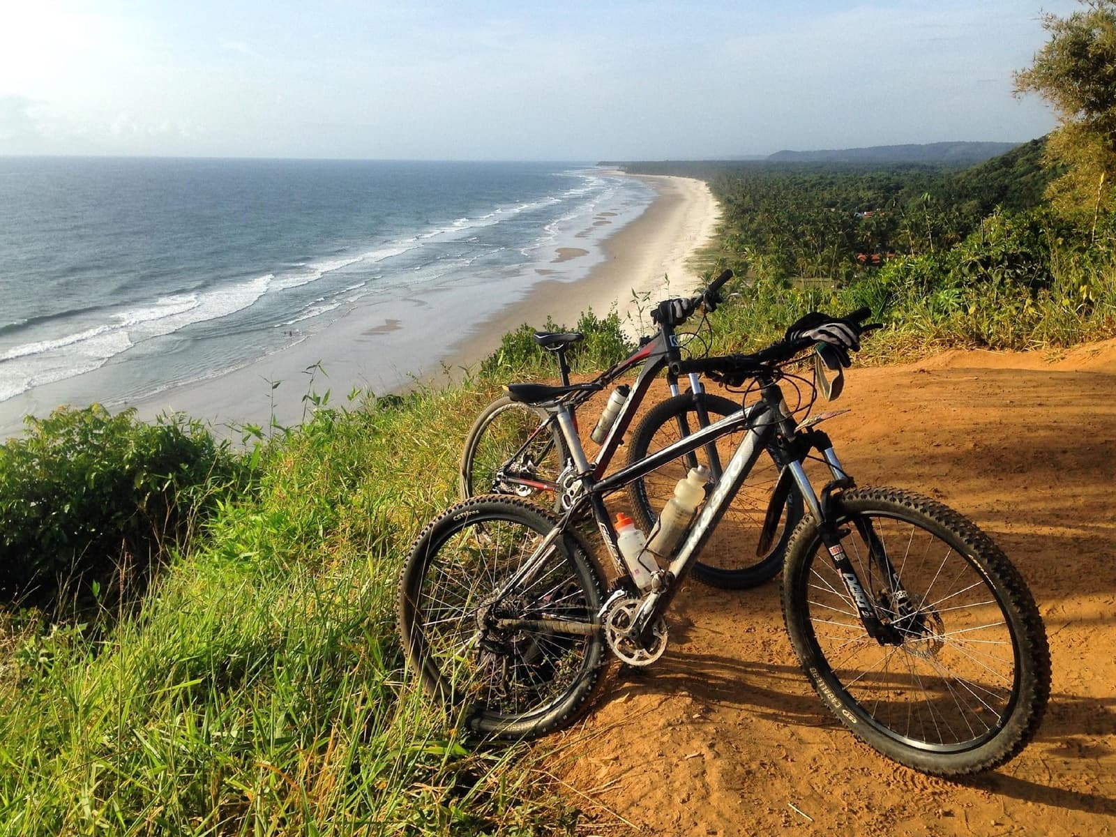 Duas bikes, uma vista, e uma praia que não tem fim. Duas mountain bikes paradas em uma trilha de terra vermelha no alto de uma falésia durante um passeio de ciclismo na Bahia, com uma longa praia deserta, ondas do Atlântico e mata atlântica costeira ao fundo