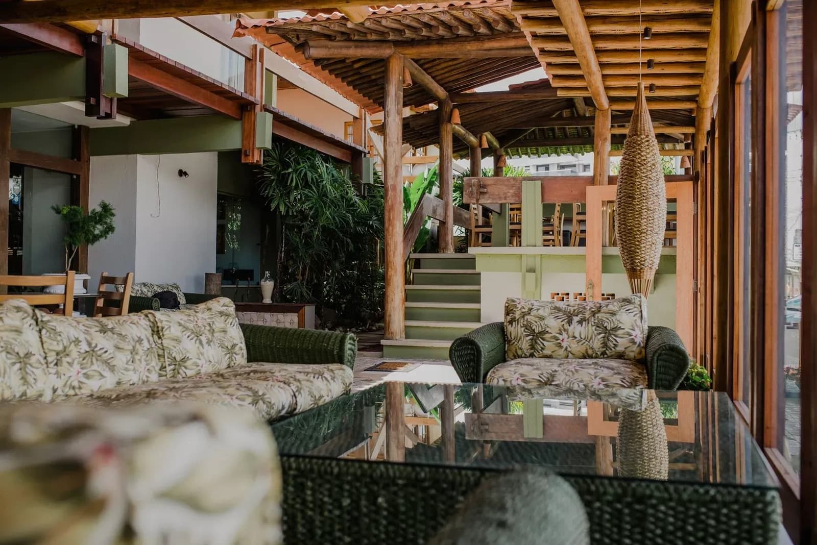 A pousada certa deixa as pernas descansadas e a cabeça leve. Lounge aberto com sofás de vime e almofadas floridas sob um telhado rústico de troncos, plantas tropicais e uma escada ao fundo em uma pousada na Rota do Cacau na Bahia de bicicleta