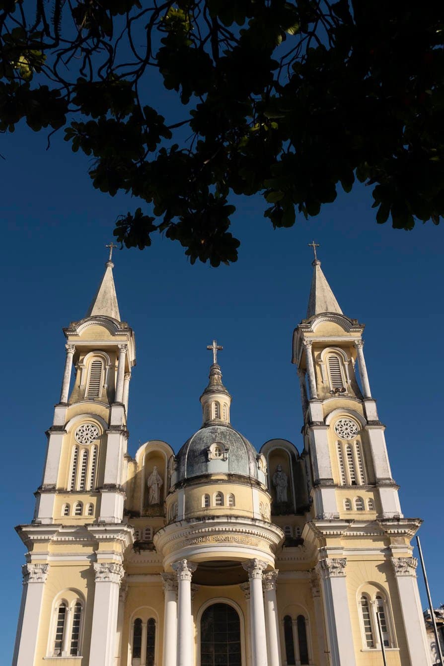 Ilhéus começa assim, com torres no céu e história escrita na pedra. Vista em ângulo baixo de uma igreja neoclássica amarelo-pálido e branca com duas torres sineiras, uma cúpula central encimada por uma cruz e a inscrição 'In Honorem Sancti Sebastiani' na fachada, emoldurada por galhos escuros contra um céu azul intenso em Ilhéus, Bahia.