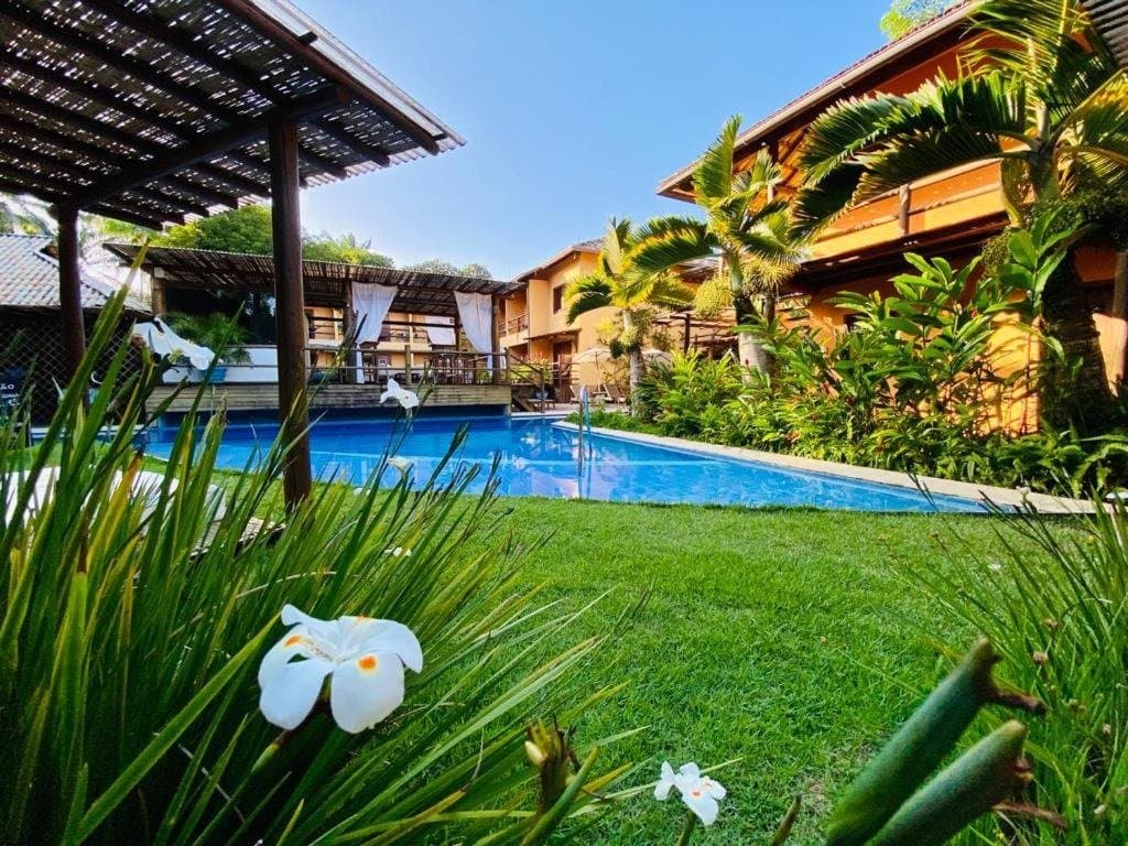 Pedalou, suou, e agora o jardim é seu por uma noite. Jardim tropical exuberante com flores brancas em primeiro plano, piscina azul e uma pousada de dois andares em tons quentes sob o céu limpo de Itacaré