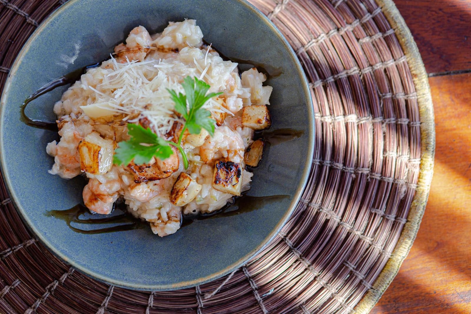 Pedalou bastante, agora é hora de comer bem. Uma tigela de cerâmica com risoto cremoso de camarão, pedaços de legume-raiz grelhado, queijo ralado, folha de salsinha fresca e fio de balsâmico, sobre um sousplat de palha trançada em mesa de madeira