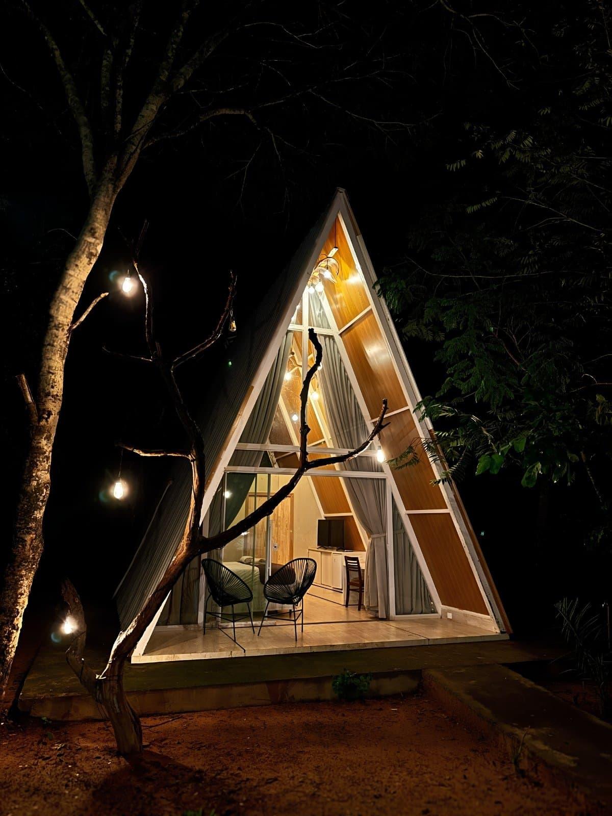 O Cerrado à noite tem esse jeito de fazer você não querer sair. Uma cabana em formato A-frame iluminada com luz quente à noite, rodeada por árvores escuras e cordões de lâmpadas Edison, com duas cadeiras no deck aberto — abrigo aconchegante para uma aventura no Jalapão