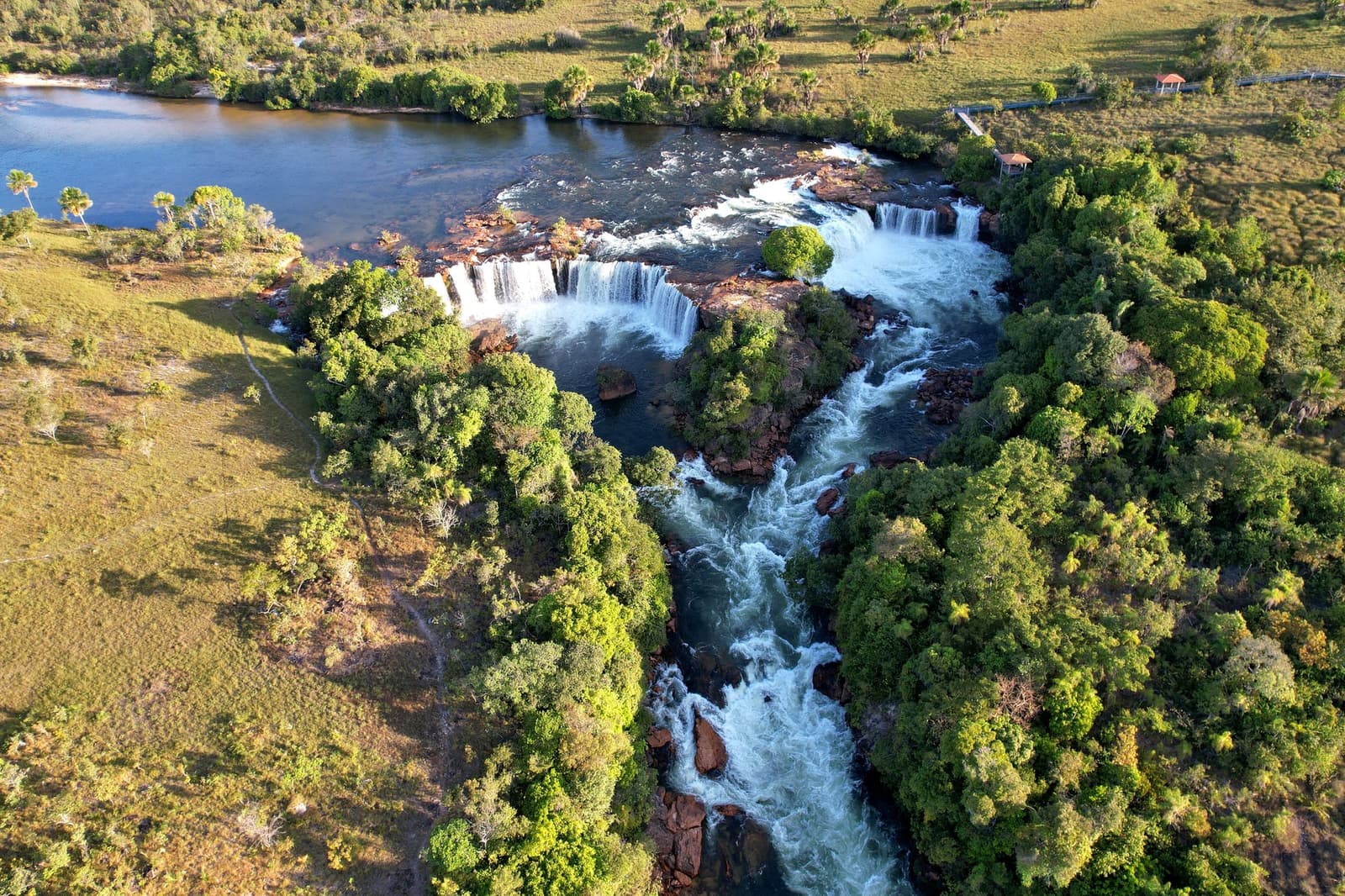 O rio não escolhe um único salto quando pode ter vários. Photo: Wirestock Vista aérea de um sistema de cachoeiras em múltiplos níveis em uma aventura no Jalapão, com corredeiras brancas despencando sobre lajes de rocha escura em um rio agitado ladeado por vegetação densa do Cerrado e campos abertos
