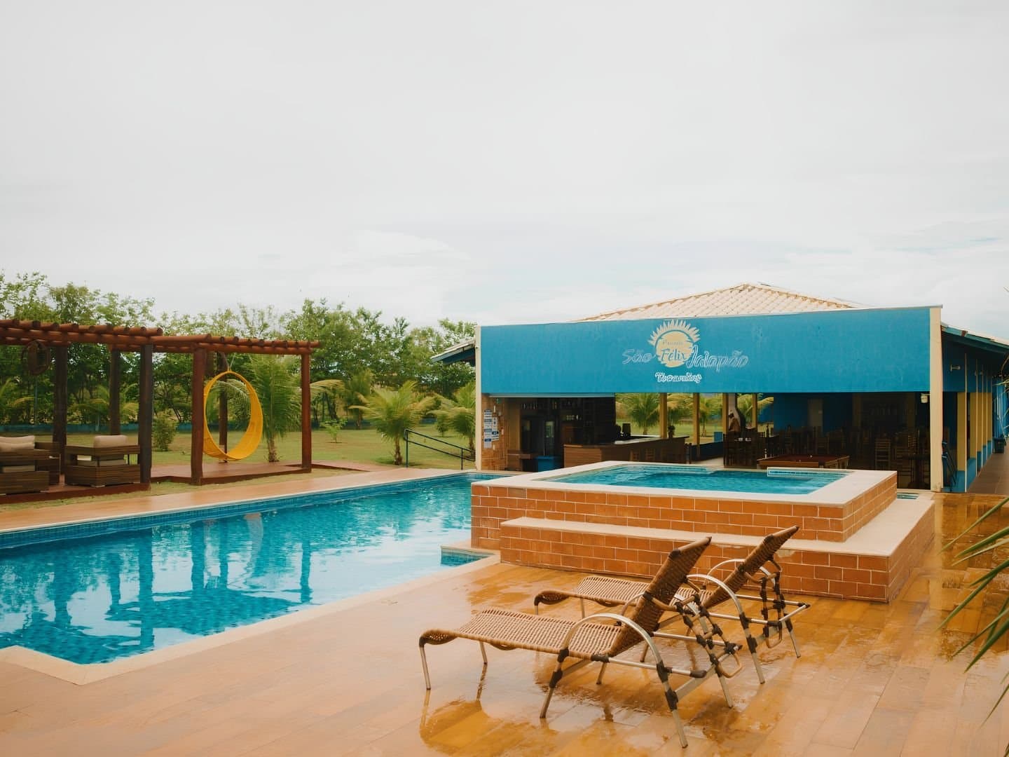 Sore legs from the dunes? The pool is already waiting for you. Outdoor pool and raised hot tub at a guesthouse in São Félix do Tocantins, with wicker sun loungers on a wet terracotta deck, a yellow hanging chair under a wooden pergola, and the teal-painted inn building in the background — a comfortable base for a jalapão trip