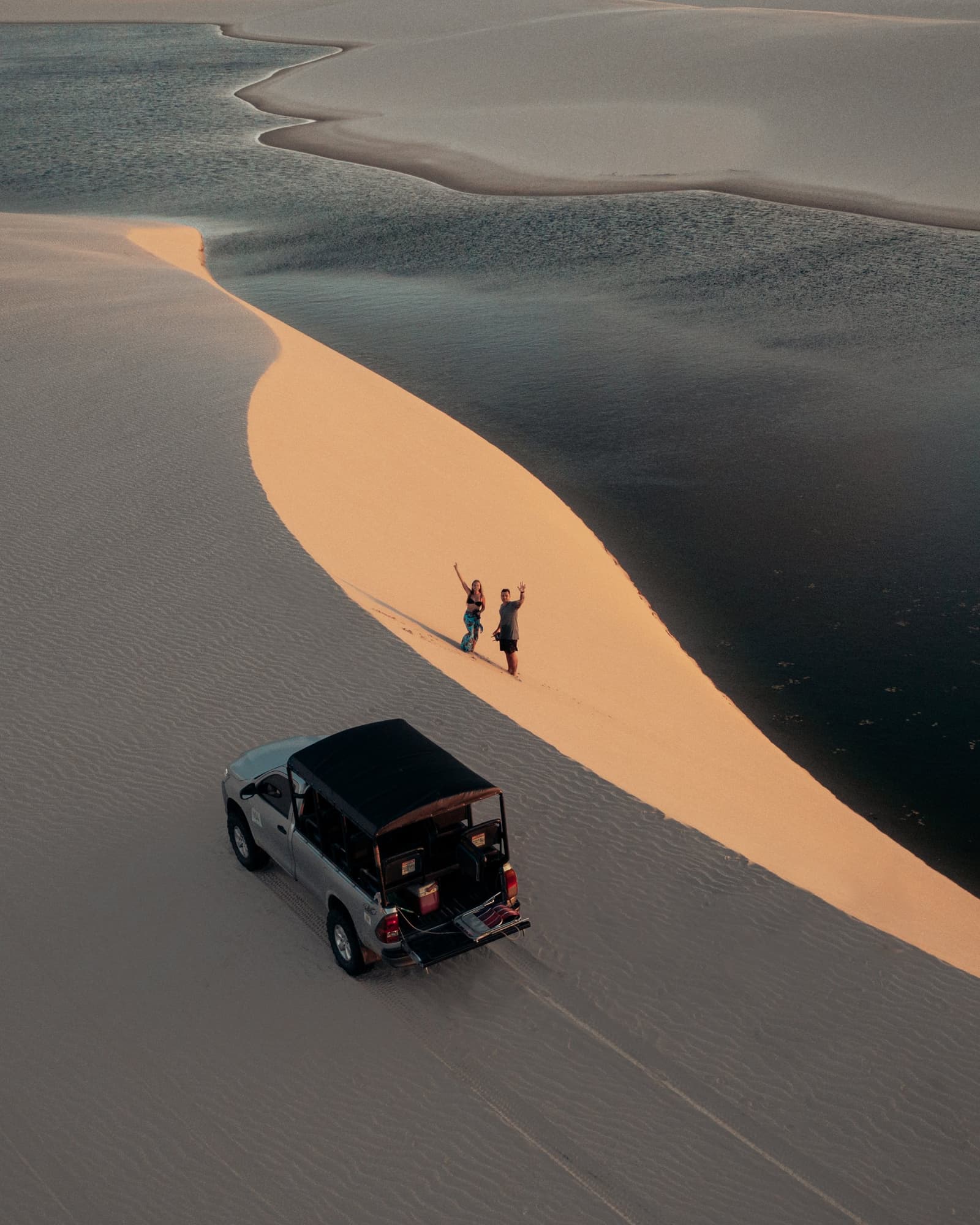 Chegaram até aqui de 4x4, mas a sensação é de ter chegado ao fim do mundo. Vista aérea de dois viajantes acenando sobre uma crista de duna dourada ao entardecer, ao lado de uma lagoa escura e reflexiva, com um 4x4 estacionado abaixo — cena do roteiro completo pelos Lençóis Maranhenses