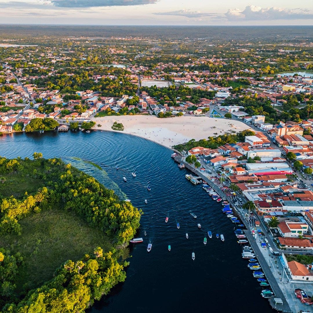 A cidade abraça o rio como quem não tem pressa de ir embora. Vista aérea de Barreirinhas mostrando o rio Preguiças contornando uma praia de areia branca, barcos coloridos atracados à beira do rio e a cidade se espalhando pela vegetação — porta de entrada para o roteiro completo dos Lençóis Maranhenses