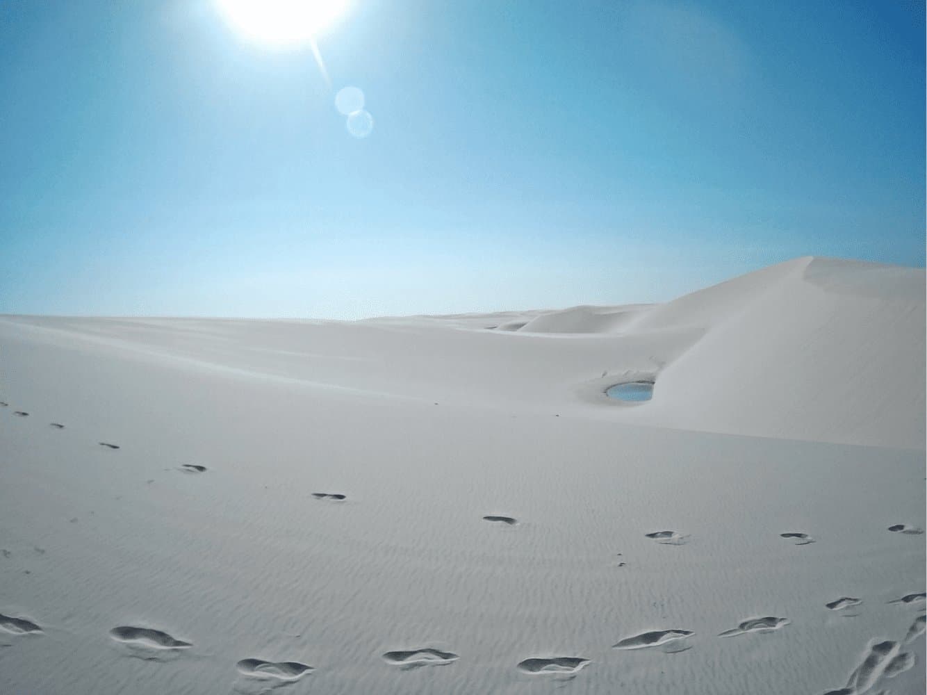 Alguém passou por aqui, encontrou água e provavelmente não quis mais sair. Pegadas descalças cruzam vastas dunas de areia branca dos Lençóis Maranhenses em direção a uma pequena lagoa azul encravada entre as cristas, sob um sol intenso