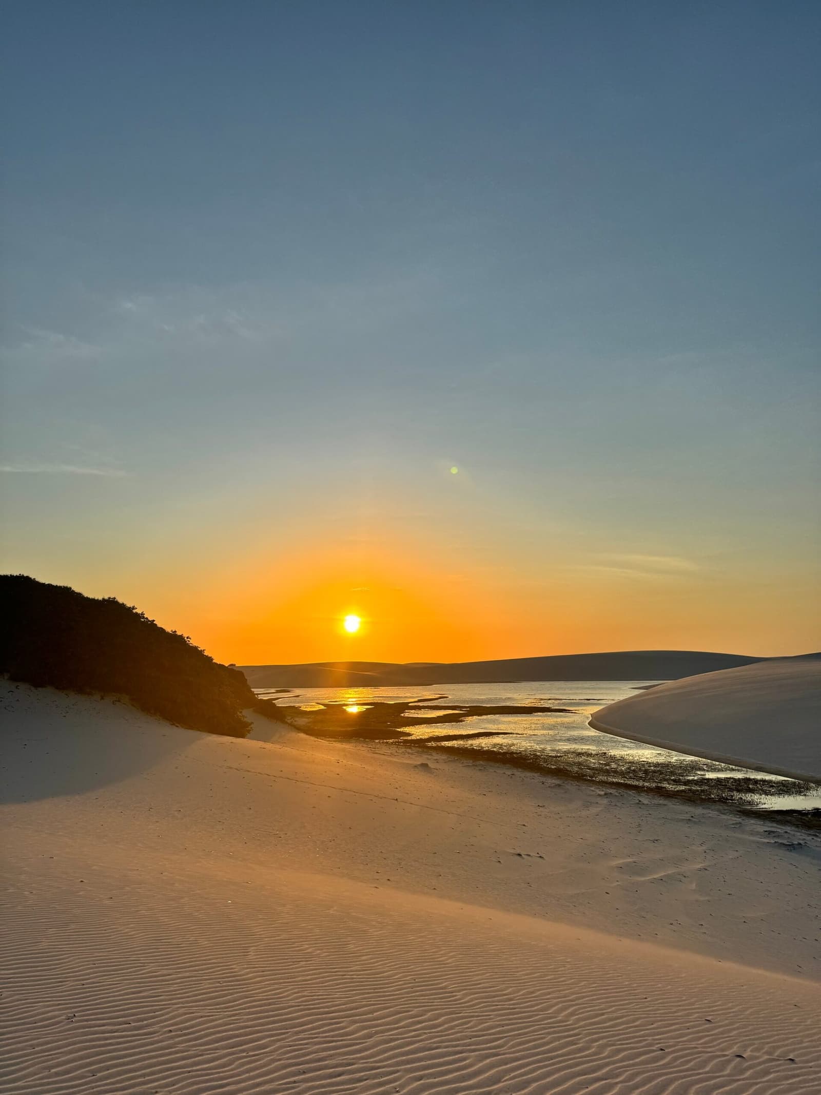 O fim do dia nos Lençóis tem aquela cor que fica na memória. Pôr do sol dourado sobre um canal de maré serpenteando entre dunas de areia branca nos Lençóis Maranhenses, refletindo luz alaranjada na água rasa