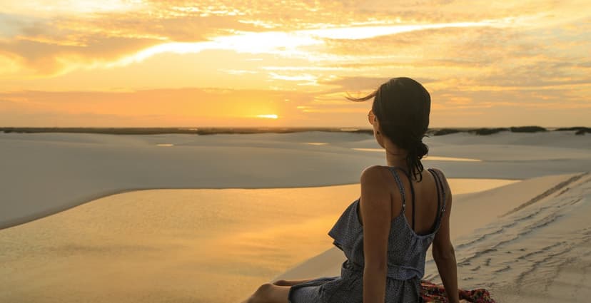 A woman sits alone on a white sand dune watching a vivid golden sunset over the Lençóis Maranhenses full itinerary landscape of dunes and shallow lagoons A woman sits alone on a white sand dune watching a vivid golden sunset over the Lençóis Maranhenses full itinerary landscape of dunes and shallow lagoons